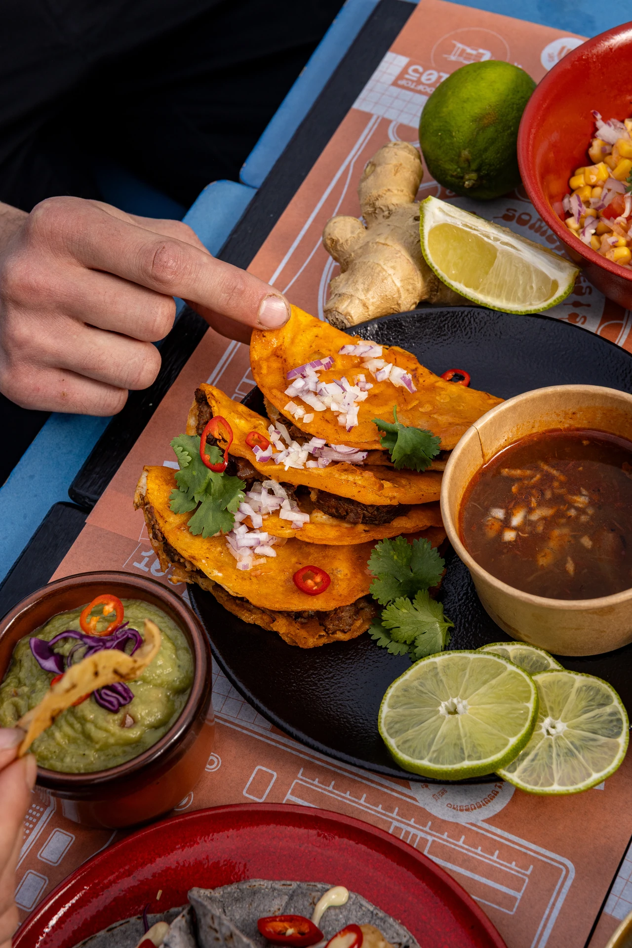 Main saisissant un taco birria orange avec consommé, guacamole et citron vert